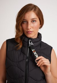 Svart, vadderad ärmlös väst med hög krage, med dragkedja fram och en logotyp på dragfliken. Slät nylon med en modern design.