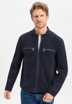 Man met lichtbruin haar en baard, gekleed in een marineblauwe jas met rits en vier zichtbare ritstaszakken en een blauwe spijkerbroek tegen een witte achtergrond.