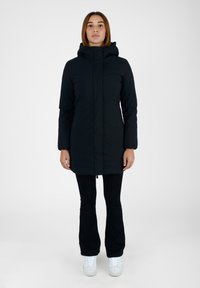 Giacca a vento nera con cappuccio e zip completa, dotata di due tasche laterali e un tessuto leggermente testurizzato. Indossata sopra pantaloni neri e scarpe bianche.