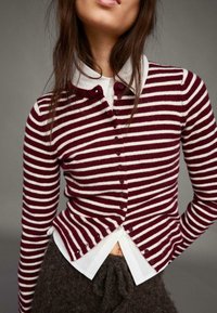 Kardigan met bordeauxrode en witte strepen, een ronde halslijn en knopen aan de voorkant. Draagt over een wit overhemd met kraag, wat gelaagde texturen laat zien.