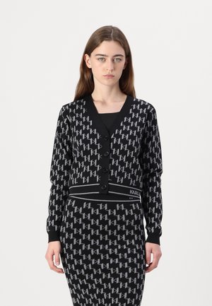 Zwarte cardigan met een geometrisch grijs patroon, voorzien van een V-hals, lange mouwen en vijf knopen. Gecombineerd met een bijpassende rok met hetzelfde patroon.