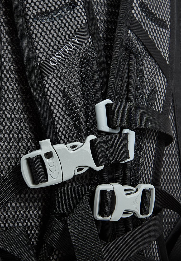 Sac à dos en mesh noir avec des boucles et des sangles en plastique gris. Marquage visible sur la sangle, avec une surface texturée et des composants ajustables.