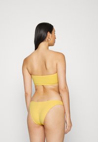 Top de bikini bandeau amarelo e parte de baixo de bikini combinando com um padrão texturizado, exibidos de uma vista traseira contra um fundo simples.