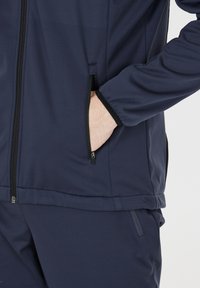 Marineblauwe sportjack met ritszakken en een gladde, rekbare textuur. Het ontwerp heeft elastische manchetten en een getailleerde taille.