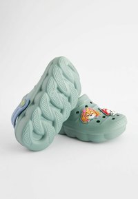 Ljust gröna träskor gjorda av flexibelt material med flera ventilationshål, prydda med söta karaktärstema dekorationer.