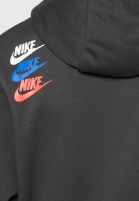 Černá mikina s kapucí, která na levém rameni zobrazuje tři loga Nike swoosh v bílé, modré a červené barvě. Měkká, texturovaná látka a ležérní design.