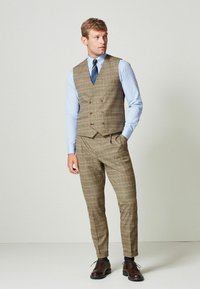 Muž oblečený v hnedoštvorcovej vestě a nohaviciach, s svetlo modrou košeľou, pruhovanou kravatou a tmavo hnedými formálnymi topánkami, stojí s jednou rukou v vrecku.