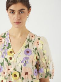 Broderad klänning i genomskinligt tyg med färgglada blommönster i nyanser av lila, gult, rosa och grönt. V-ringad design och puffärmar.