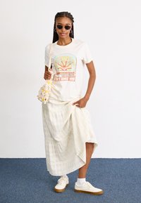 Graphic t-shirt in crème met een kleurrijke print, gecombineerd met een lichte, gestructureerde, geruite rok en witte sneakers. Houdt een gehaakte tas met bloemaccenten vast.