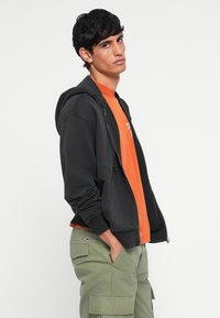 Sort zip-up hættejakke med snoretræk, over en orange t-shirt. Kombineret med grønne cargo-bukser med sidelommer og logodetaljer.
