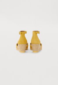 Sandales compensées en suède jaune avec une bride à la cheville, détails tissés texturés sur le talon, dos fermé et accent de boucle circulaire lisse.