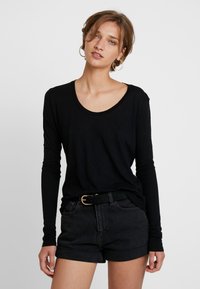 Camisa preta de manga longa com decote em forma de U, combinada com calções pretos de cintura alta. Detalhes notáveis incluem um cinto preto texturizado com uma fivela dourada.