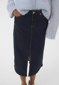 Dunkler Denim-Midirock mit einem vorderen Schlitz, gelben Nähten und figurbetontem Schnitt. Kombiniert mit einem hellblauen, flauschigen Strickpullover.