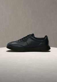 Baskets basses en cuir noir avec lacets, exposées sur une surface claire contre un fond neutre.