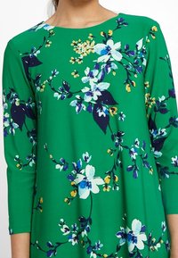 Green floral dress featuring a round neckline, three-quarter sleeves, and navy, white, and yellow floral patterns throughout. Made from soft fabric.