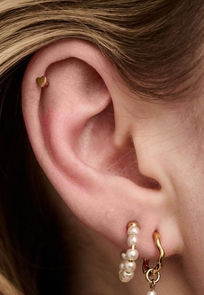 Goldene Herz-Stift-Ohrringe im oberen Ohrläppchen, kombiniert mit einem goldenen und Perlen-Hoop-Ohrring im unteren Ohrläppchen; die Haut ist hell.