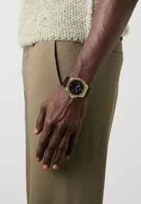 Reloj negro y dorado con una esfera redonda, correa de silicona negra y diseño minimalista; llevado en una mano sobre una tela de pantalón verde oliva.