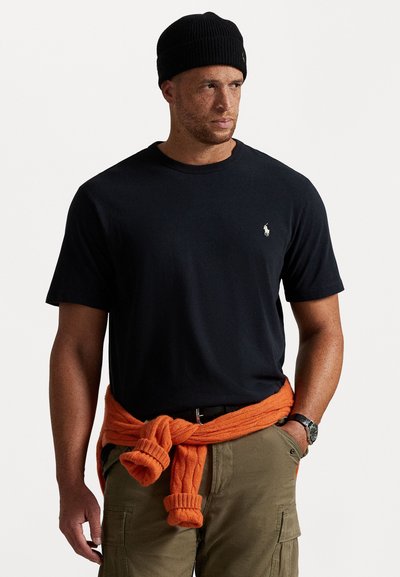 Melnas īsām piedurknēm T-krekls ar mazu izsietu logo, pārī ar olīvzaļa krāsas kargo biksēm un oranžu adītu džemperi, kas sasietas ap vidukli.