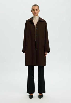 Femme portant un manteau marron foncé boutonné par-dessus un col roulé beige, un pantalon noir et des chaussures noires, debout devant un fond clair uni.
