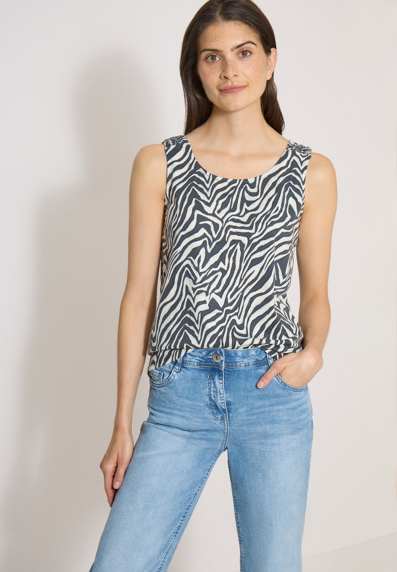 Femme aux longs cheveux foncés portant un haut sans manches à imprimé zèbre noir et blanc et un jean bleu clair, une main dans la poche.