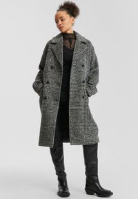 Grijs en zwart houndstooth-patroon wollen overjas met een relaxte pasvorm, dubbele knopen en grote revers. Gecombineerd met kniehoge zwarte laarzen.