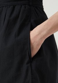 Vestido negro de lino con cintura fruncida y bolsillos laterales, con textura visible y acabado suave. Mano situada en un bolsillo.
