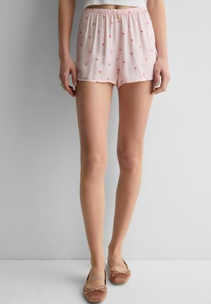 Kobieta ubrana w jasnoróżowe szorty z drobnymi nadrukami wiśni, biały krótki top i brązowe baleriny, stojąca na jednolitym tle.