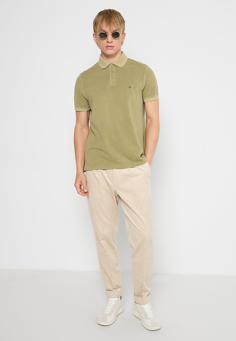 Polo de color verde oliva con cuello, mangas cortas y un sutil logo en el pecho. Combinado con pantalones de pana beige y zapatillas blancas.