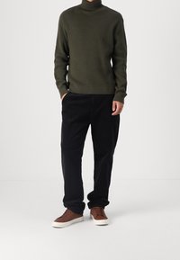 Pull à col roulé en maille vert foncé, pantalon en velours côtelé noir et baskets marron à lacets avec une semelle blanche. Tissu texturé et coupe décontractée.