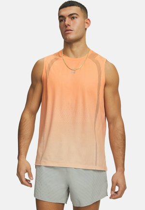 Gespiierde man die een mouwloos oranje sportshirt en lichtgrijze korte broek draagt, staand tegen een witte achtergrond, licht naar links kijkend.