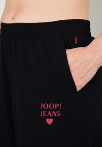 Mustat verkkarit, joissa on joustava vyötärö, punaisella printatulla "JOOP! JEANS" -logolla ja sydänyksityiskohdalla. Oikealla puolella näkyy sivutasku.