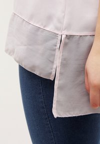 Lyserød bluse med et gennemsigtigt nederste lag; har en lige skæring med sideslidser og en glat tekstur, båret over mørk denim.