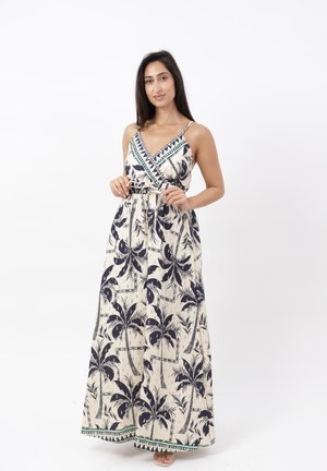 Vrouw die riem aanpast van een mouwloze maxi-jurk met zwart palmboomprint en geometrische bies op een crèmekleurige achtergrond, staand op een witte achtergrond.