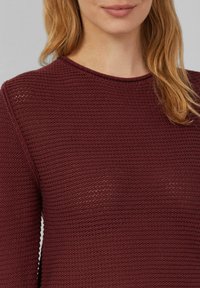 Vrouw in een wijnrood gebreid sweater met een ronde halslijn, weergegeven van schouders tot lippen, met blond haar over haar rechter schouder.