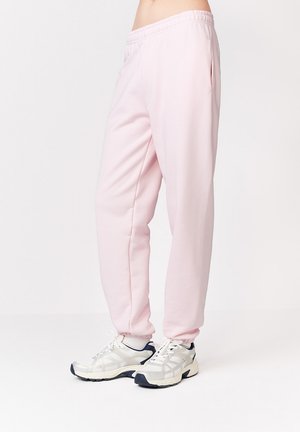 Osoba nosząca jasnoróżowe spodnie dresowe z elastycznym pasem i białe buty sportowe na jednolitym tle.