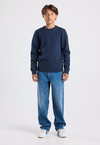 Tummansininen neule, jossa on pyöreä kaula-aukko, ribbireunat hihoissa ja helmassa; yhdistetty vaaleansinisiin rentoihin farkkuihin ja valkoisiin urheilukenkiin, joissa on sinisiä yksityiskohtia.