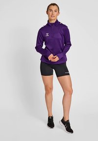 Veste de sport violette à fermeture éclair quart, manches longues, avec détail de logo. Associée à un short noir et des chaussures de sport noires.