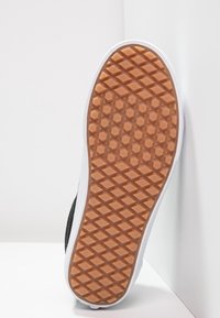 Semelle de sneaker avec un motif en caoutchouc brun texturé, bordée d'une semelle intermédiaire blanche. Le côté présente un matériau supérieur noir minimal.