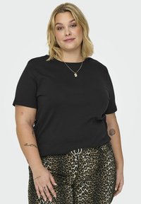 T-shirt preto de manga curta num corte descontraído, combinado com calças de padrão de leopardo. Detalhes notáveis incluem um decote redondo e tatuagens visíveis.
