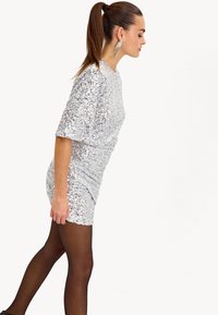 Zilveren paillettenjurk met korte mouwen, getailleerd ontwerp en een gestructureerd oppervlak. Model draagt zwarte panty’s en lange, hangende oorbellen.