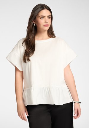 Mujer con cabello largo y castaño viste una blusa blanca de manga corta con volantes en el bajo y pantalones negros, posando frente a un fondo claro.