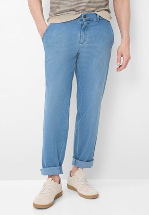 Uomo che indossa jeans azzurri con risvolti, sneakers beige e un maglione beige lavorato a maglia con le mani nelle tasche.