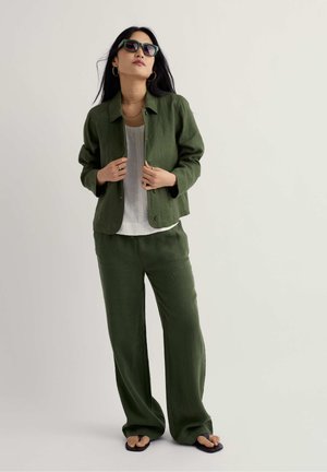 Žena v tmavozelenom ľanovom obleku s nohavicami širokých nohavíc, bielym topom, čiernymi sandálmi, slnečnými okuliarmi a zlatými kruhovými náušnicami na jednoliatom pozadí.