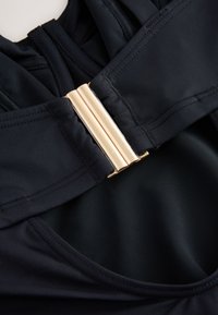 Gros plan d’un maillot de bain en tissu noir avec une large bretelle et une boucle rectangulaire dorée brillante à l’arrière.