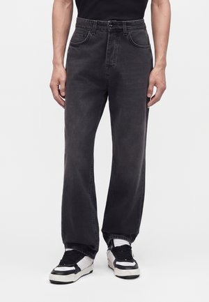 Person iført løstsiddende sorte jeans og sorte og hvide sneakers, stående mod en ensfarvet baggrund.