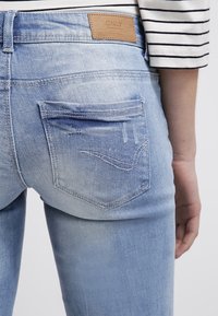 Světle modré denimové džíny s opotřebovaným vzhledem, obsahující dvě zadní kapsy a hnědou koženou nášivku na pase.