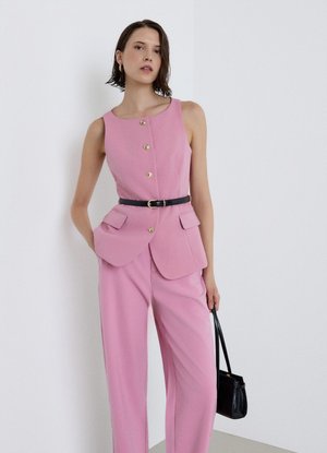 Donna con gilet rosa senza maniche con bottoni dorati e pantaloni coordinati, che tiene una borsa nera, su uno sfondo bianco semplice.