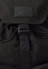 Juodas kuprinis krepšys su tekstūruota nailono išore. Įrengtas logotipo užtrauktuku, reguliuojamu diržu ir saugiu uždarymu.