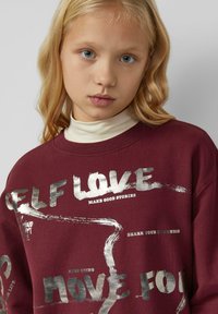 Bordeauxrotem Sweatshirt mit silberner Druckschrift. Mit Rundhals, langen Ärmeln und weichem Material. Der Text enthält motivierende Sprüche.