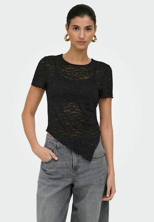 Kobieta w czarnej koronkowej asymetrycznej bluzce na czarnym biustonoszu, zestawiona z szarymi jeansami z wysokim stanem i złotymi kolczykami kołami, stojąca z ręką w kieszeni.
